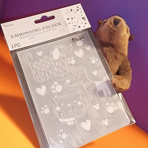 May include: Clear embossing folder with dog designs, hearts, and paw prints. The package reads "Darice EMBOSSING FOLDER" and "1PC". A brown stuffed animal is partially visible in the background.