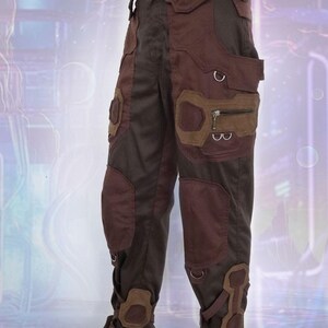 K&ouml;nnte beinhalten: Braune und schwarze Cargohose mit mehreren Taschen und Riemen. Die Hose hat ein einzigartiges Design mit einer Mischung aus Texturen und Materialien.