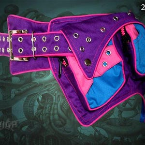 May include: A purple and pink fabric belt with a blue pocket and multiple silver eyelets. The belt has a large silver buckle and a pink trim.