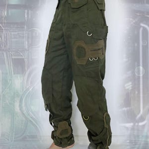 K&ouml;nnte beinhalten: Gr&uuml;ne Cargohose mit mehreren Taschen und Riemen. Die Hose hat ein einzigartiges Design mit einem gro&szlig;en Patch am rechten Oberschenkel und Riemen um die Kn&ouml;chel.