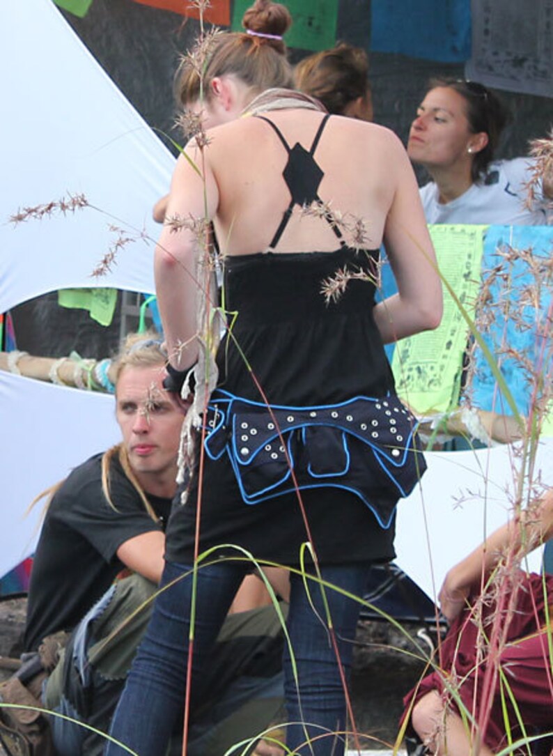 May include: A woman wearing a black tank top with a black and blue studded belt. She is standing in front of a white background with plants in the foreground.