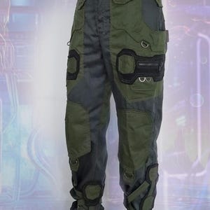 K&ouml;nnte beinhalten: Gr&uuml;n-schwarze Cargohose mit mehreren Taschen und Riemen. Die Hose hat ein futuristisches Design mit verst&auml;rkten Knien und Kn&ouml;cheln.