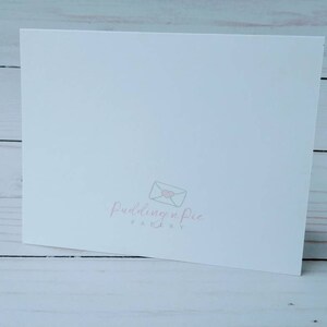 May include: A white folded card with the logo "Pudding n Pie Paperie" in light pink. The logo features an envelope with a heart. The card is on a light wood surface.