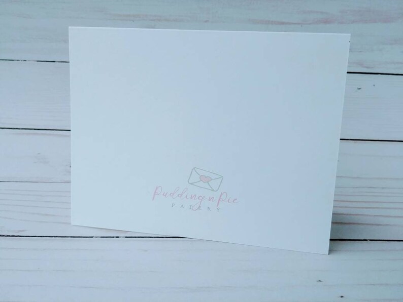 May include: A white folded card with the logo "Pudding n Pie Paperie" in light pink. The logo features an envelope with a heart. The card is on a light wood surface.