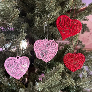 K&ouml;nnte beinhalten: Vier herzf&ouml;rmige Ornamente mit filigranen Spitzenmustern. Zwei sind rot, eines ist rosa und eines ist in einem helleren Rosaton. Die Ornamente h&auml;ngen an einem gr&uuml;nen Weihnachtsbaum.