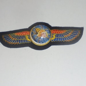 May include: A black felt winged patch with a gold trim and a round, colorful, glass center featuring a woman's face and a spider web.