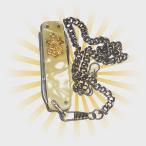 May include: A folding pocket knife with a cream-colored pearlescent handle featuring a gold-toned emblem of the Papal tiara and crossed keys. The knife is attached to a dark metal link chain with a spring-loaded clasp at the bottom end.