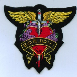 May include: Embroidered patch featuring a red heart pierced by a sword, with gold wings and the name "BON JOVI" in gold lettering. The patch has a black background and purple floral accents.