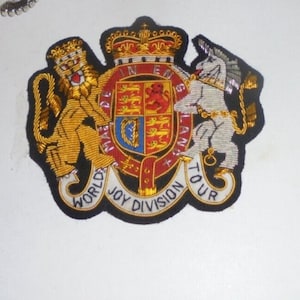 May include: Embroidered patch featuring a royal crest design with a lion and unicorn flanking a shield. The crest is in gold, red, and blue, with the text "WORLD JOY DIVISION TOUR" in white. The patch has a black border.