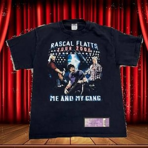May include: Black t-shirt featuring a graphic of Rascal Flatts with the text "Rascal Flatts Tour 2006" and "Me and My Gang". The shirt has a concert ticket attached to the bottom.