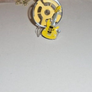 May include: A yellow electric guitar pendant necklace. The guitar is on a silver stand and has a silver chain.