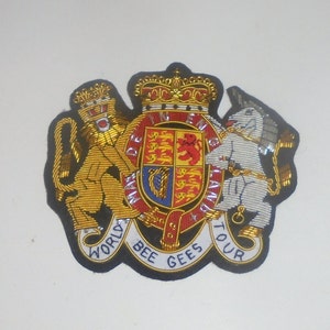 May include: A gold and white embroidered patch featuring a lion, a unicorn, and a coat of arms with the text "WORLD BEE GEES TOUR".