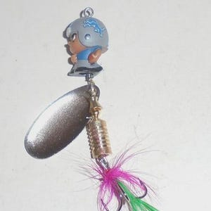 May include: A fishing lure featuring a Detroit Lions football player figurine on top. The lure has a silver blade, a gold-colored cylindrical body, and a hook adorned with pink and green feathers. The figurine wears a gray helmet.