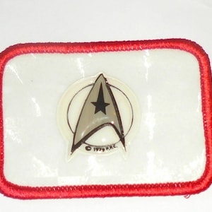 May include: A red-bordered white patch with a silver Star Trek insignia featuring a stylized starship and a five-pointed star. The insignia is surrounded by a white circle with a black outline. The text "© 1979  NCC" is visible below the insignia.