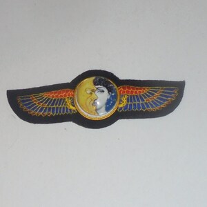 May include: A black fabric patch with a gold-trimmed circle featuring a stylized image of a face with a sun and moon design. The circle is surrounded by a pair of red, blue, and gold wings.