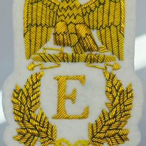 May include: A white embroidered patch with a gold eagle, a gold letter E, and gold laurel wreaths. The eagle is perched on a gold banner.