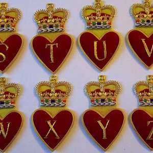 May include: Eight embroidered felt heart-shaped patches with gold crowns and letters S through Z. The hearts are red with gold trim and the crowns are gold with red, green, and white accents.