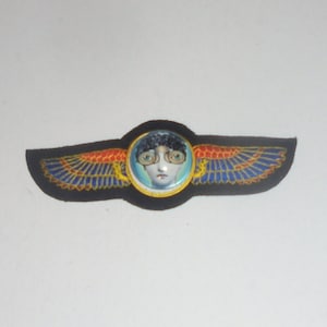 May include: A black fabric patch with a gold trim and a red, blue, and yellow embroidered design. The center of the patch features a round, clear glass dome with a picture of a person with blue eyes and black hair wearing glasses.