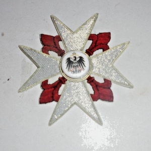 May include: A decorative cross-shaped emblem with a central circular design featuring a black eagle. The cross has silver glittered arms, red accents, and gold trim. The emblem is likely a badge or a decorative element.