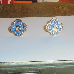 May include: Two gold-toned, four-leaf clover-shaped pins with a light blue enamel center. Each pin features a detailed design in the center, surrounded by a gold border. The pins are displayed on a white surface.