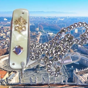 May include: A pocket knife with a pearlescent white handle featuring a gold-toned emblem of the Papal tiara and keys above a small shield-shaped crest. A silver-toned metal chain with a spring-loaded clasp is attached to the base of the handle.