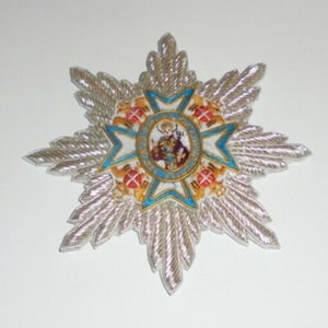 May include: A silver star-shaped badge with a blue and white center featuring a religious figure in a gold frame. The star has a silver fringe around the edges.