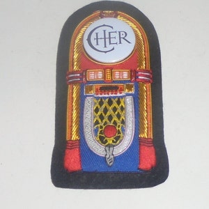 May include: Embroidered patch in the shape of a jukebox, featuring the name "CHER" in a white circle. The design includes red, blue, gold, and black accents, with a detailed, textured appearance. The patch has a black border.