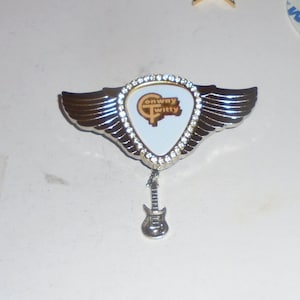 May include: A silver-toned guitar pick-shaped pin with the words "Conway Twitty" in a gold color. The pin is surrounded by a ring of clear rhinestones and has silver wings. A small silver guitar charm hangs below.
