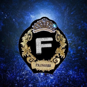 May include: A black velvet patch with a gold embroidered crown and a silver letter F adorned with rhinestones. The patch also features gold embroidered leaves and the word "Princess" in gold lettering.