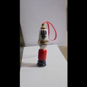 May include: A small wooden figurine of a person wearing a red and black uniform with a white porcelain hat featuring a gold eagle design. The hat has a red ribbon attached.
