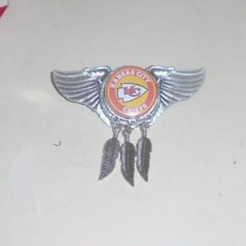 May include: Silver Kansas City Chiefs pin with wings and feathers. The pin features the team's logo with the words "Kansas City Chiefs" and a red arrow pointing to the right.