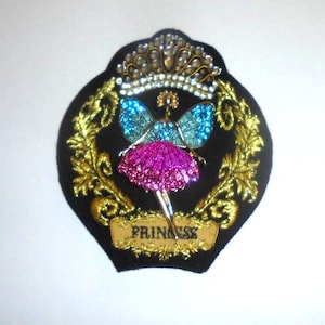 May include: A black fabric patch with gold embroidery depicting a fairy with blue wings and a pink skirt. The fairy is wearing a crown and the patch says "Princess" in gold lettering.
