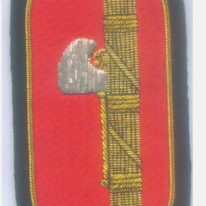 May include: A red embroidered patch with a gold and silver design. The design features a stylized eagle with a rope and a bundle of sticks.