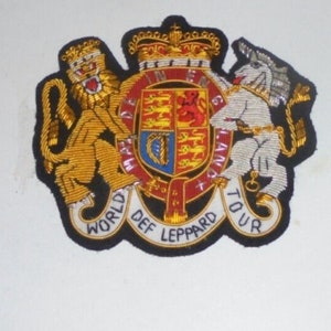 May include: A black and gold embroidered patch with a lion and unicorn on either side of a royal coat of arms. The patch has the text "World Def Leppard Tour" around the outside.