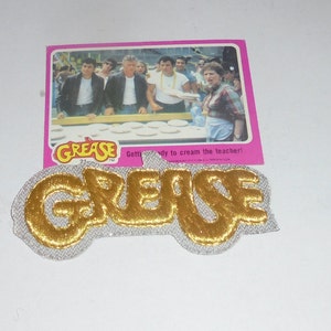 May include: A gold-colored "Grease" patch with a white background. The patch is embroidered and has a slightly raised texture. The text "Grease" is in a stylized font. The patch is accompanied by a pink card with a picture of the cast of the movie "Grease" and the text "Get ready to cream the teacher!"