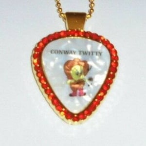May include: A gold guitar pick necklace with a white background and red rhinestones around the edge. The pick features a cartoon image of Conway Twitty and the text "Conway Twitty".