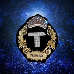 May include: A black and gold embroidered patch with a crown, the letter "T" adorned with rhinestones, and the word "Princess" below. The patch is set against a dark blue background with a blurry, abstract design.