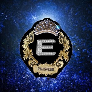 May include: A black emblem with a gold crown and laurel wreath design. The letter "E" is encrusted with rhinestones. The word "PRINCESS" is written on a banner below the letter. The background is a dark blue.