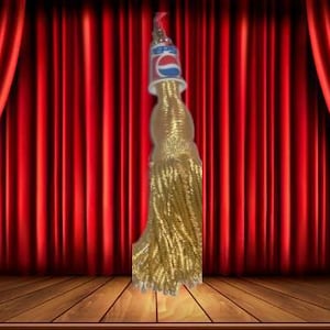 May include: A gold tassel with a Pepsi logo on top stands on a wooden stage in front of red curtains. The tassel is thick and ornate, with a long, flowing design.