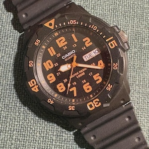 May include: A black Casio quartz watch with a textured resin strap. The round dial features orange Arabic numerals, a 24-hour inner ring, and a day-date window at the 3 o'clock position. The bezel is marked with orange numbers up to 55.