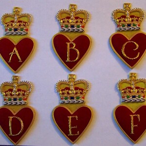 May include: Six embroidered patches featuring a red heart shape with a gold border, each with a different letter from A to F in gold. Each heart is topped with a gold crown with colorful jewels. The patches are on a yellow background.