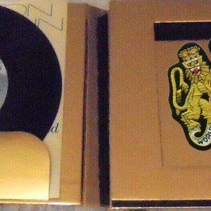 May include: A gold-colored box with a black vinyl record inside. The box has a gold-colored patch with a lion and a unicorn, and the text "World Elton John Tour".