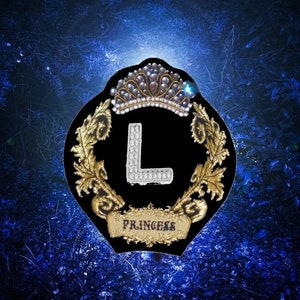 May include: A black emblem with a gold crown and laurel wreath design, featuring the letter "L" in silver rhinestones. The word "PRINCESS" is displayed on a gold banner. The background is a dark blue, leafy pattern.