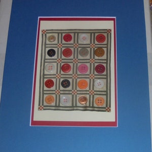 May include: A framed artwork featuring a quilt made of various colored buttons arranged in a grid pattern. The quilt is framed in a red and white mat and a blue frame.