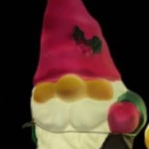 May include: A whimsical garden gnome figurine with a red hat, white beard, and green accents. The gnome holds a lantern and a red apple. The figurine is illuminated, suggesting it may be a decorative light.