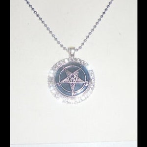 May include: A silver necklace with a round pendant featuring a black pentagram with a white star inside. The pendant is surrounded by a circle of clear rhinestones.