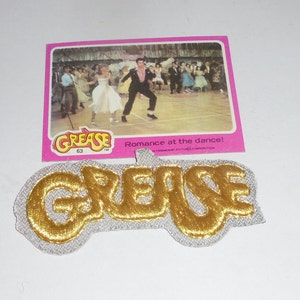 May include: A gold-colored "Grease" patch with a white background. The patch is a popular collectible from the 1978 movie "Grease".