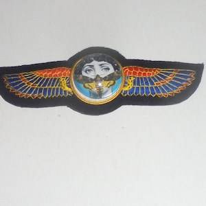 May include: A black fabric patch with embroidered wings in red, blue, and gold. A round, colorful button with a woman's face and a moth is in the center.