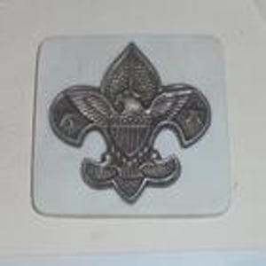 May include: A silver-colored metal belt buckle featuring the Boy Scouts of America emblem, a fleur-de-lis with an eagle and shield design. The buckle is rectangular with rounded corners and is set against a white background.