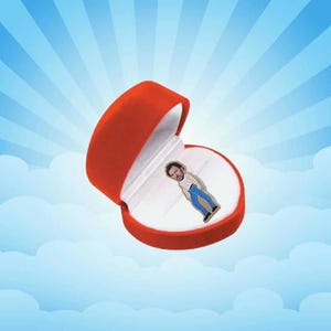 May include: A red velvet ring box with a white interior holds a small pin of a person wearing blue jeans and a white shirt. The background is a blue sky with white clouds.